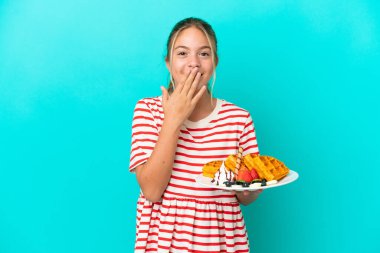 Mavi arka planda izole edilmiş waffle tutan küçük beyaz kız mutlu ve gülümsüyor.