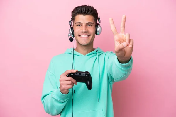 若いですハンサムな白人男性遊びますとともにAビデオゲームコントローラ孤立ピンクの背景笑顔と勝利サインを示します — ストック写真