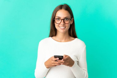 İzole edilmiş mavi arka planda genç bir kadın kameraya bakıyor ve cep telefonunu kullanırken gülümsüyor.