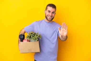 Genç Kafkasyalı, sarı arka planda izole edilmiş eşyalarla dolu bir kutuyu eline aldığında mutlu bir ifadeyle selam veriyor.