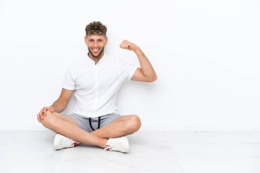 Genç sarışın adam beyaz arka planda izole bir şekilde yerde oturuyor. Güçlü bir hareket yapıyor.