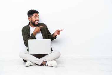 Yerde dizüstü bilgisayarı olan genç Brezilyalı bir adam beyaz parmakla işaret ederek bir ürün sunuyor.