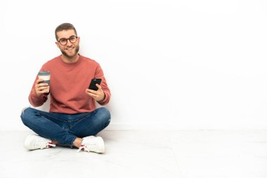 Yerde oturan yakışıklı genç bir adam elinde kahve ve bir cep telefonu tutuyor.