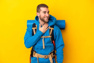 Young caucasian mountaineer man with a big backpack isolated on yellow background looking up while smiling