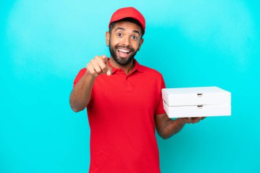 İş üniformalı Brezilyalı bir adam mavi arka planda izole edilmiş pizza kutularını topluyor. Şaşırmış ve ön tarafı işaret ediyor.