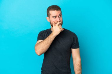 Mavi arka planda soyutlanmış yakışıklı beyaz bir adam. Şüpheleri var ve kafası karışmış bir yüz ifadesi var.