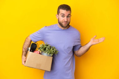 Genç Kafkasyalı, sarı arka planda izole edilmiş eşyalarla dolu bir kutuyu tutarken el kaldırırken şüpheye düşüyor.