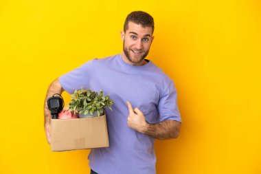 Genç Kafkasyalı, sarı arka planda izole edilmiş, sürpriz bir yüz ifadesiyle bir kutu dolusu şey alırken hamle yapıyor.