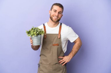 Bahçıvan beyaz bir adam sarı arka planda izole edilmiş bir bitki tutuyor. Kolları kalçasında ve gülümsüyor.