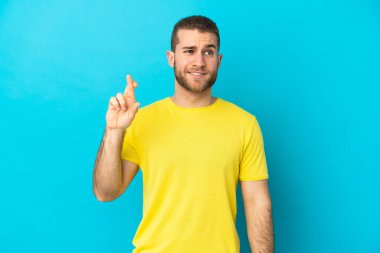 Mavi arka planda izole edilmiş genç, yakışıklı beyaz bir adam parmaklarını çapraz yapıp en iyisini diliyor.