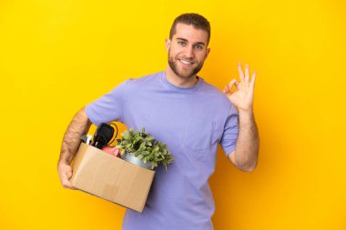 Genç Kafkasyalı, sarı arka planda izole edilmiş eşyalarla dolu bir kutuyu alırken hareket ediyor.