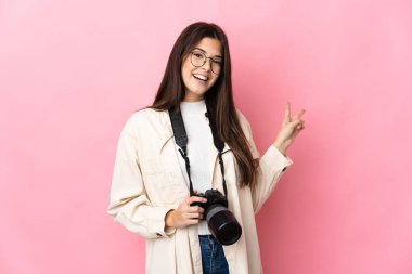 Genç fotoğrafçı Brezilyalı kız pembe arka planda izole edilmiş gülümsüyor ve zafer işareti gösteriyor.