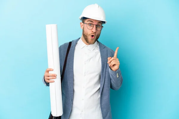 Young architect man with helmet and holding blueprints over isolated background intending to realizes the solution while lifting a finger up