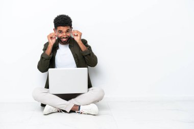 Yerde dizüstü bilgisayarı olan genç Brezilyalı bir adam gözlüklü, beyaz tenli ve şaşırmış bir şekilde oturuyor.