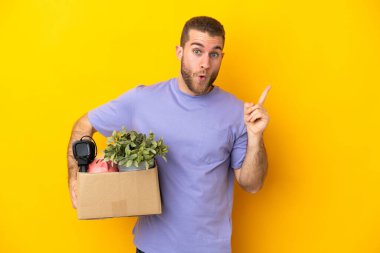 Genç Kafkasyalı, sarı arka planda izole edilmiş şeylerle dolu bir kutuyu kaldırırken bir parmağını kaldırarak çözümü bulmayı umuyor.