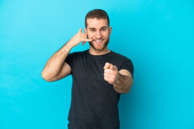 Genç yakışıklı beyaz adam mavi arka planda izole edilmiş telefon hareketleri yapıyor ve ön tarafı işaret ediyor.