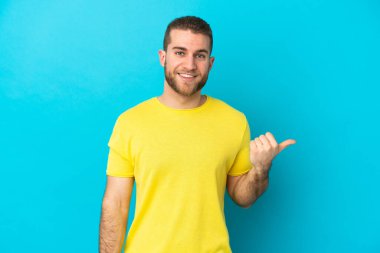 Mavi arka planda izole edilmiş yakışıklı beyaz adam bir ürünü sunmak için kenarı işaret ediyor.