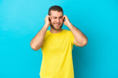 Mavi arka planda izole edilmiş yakışıklı beyaz adam hayal kırıklığına uğramış ve kulaklarını kapatmış.