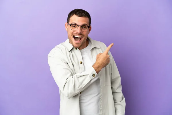 Brazilian man over isolated purple background surprised and pointing side