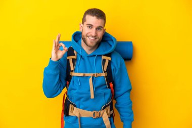 Young caucasian mountaineer man with a big backpack isolated on yellow background showing ok sign with fingers