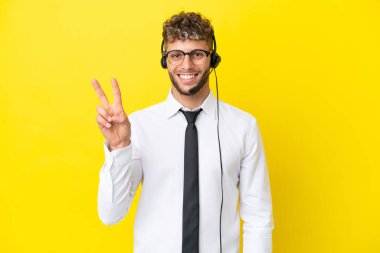 Telemarketer blonde man working with a headset isolated on yellow background smiling and showing victory sign