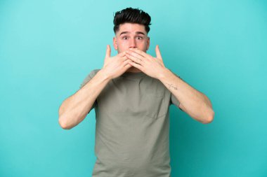 Mavi arka planda izole edilmiş genç beyaz adam elleriyle ağzını kapatıyor.