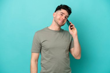 Genç beyaz adam mavi arka planda izole edilmiş cep telefonuyla sohbet ediyor.