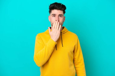 Genç beyaz adam mavi arka planda izole edilmiş, ağzı elle kapatılmış.