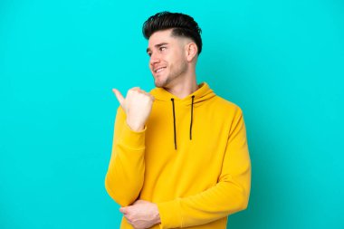 Mavi arka planda izole edilmiş genç beyaz adam bir ürünü sunmak için kenarı işaret ediyor.