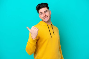 Mavi arka planda izole edilmiş genç beyaz adam bir ürünü sunmak için kenarı işaret ediyor.