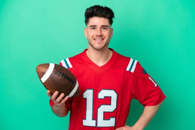 Genç beyaz adam ragbi oynuyor. Yeşil arka planda izole edilmiş. Kolları kalçasında ve gülümsüyor.