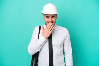 Kasklı ve elinde mavi arka planda izole edilmiş planları olan genç bir beyaz adam mutlu ve gülümseyen elleriyle ağzını kapatıyor.