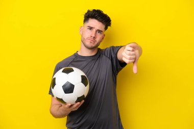 Yakışıklı genç futbolcu, sarı arka planda izole edilmiş, başparmağını negatif ifadeyle aşağı gösteriyor.