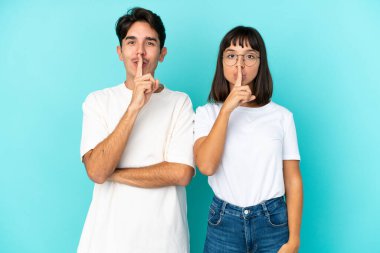 Mavi arka planda izole edilmiş genç bir melez çift. Ağız kapama ve sessizlik işareti gösteriyor.