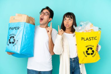 Mavi arka planda izole edilmiş plastik ve kâğıt dolu bir çanta taşıyan genç çift, bir fikrin parmağı yukarı kaldırdığını düşünüyor.