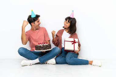 Genç melez çift, beyaz arka planda izole bir şekilde yerde oturmuş doğum gününü kutluyor ve galibiyeti kutluyorlar.