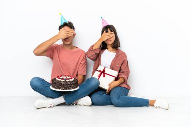 Genç melez çift, beyaz arka planda, elleri ve gözleri kapalı bir şekilde yerde oturmuş doğum gününü kutluyorlar. Bir şey görmek istemiyorum.