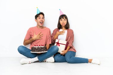 Genç melez bir çift, beyaz arka planda izole bir şekilde yerde oturmuş doğumgününü kutluyor. Şaşırmış ve şaşırmış bir şekilde sağa bakıyor.