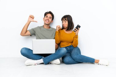 Dizüstü bilgisayarı ve cep telefonu olan genç bir çift yerde oturmuş kendini beğenmiş ve gururlu bir şekilde aşk konseptini benimsiyor.