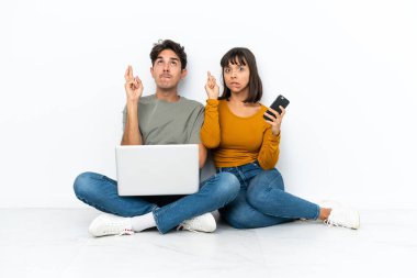 Dizüstü bilgisayarı ve cep telefonu olan genç çift yerde oturuyor ve en iyisini diliyorlar.