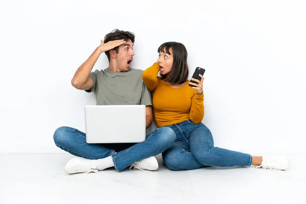 Young couple with a laptop and mobile sitting on the floor with surprise and shocked facial expression