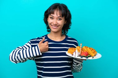 Elinde waffle tutan genç Arjantinli kadın mavi arka planda izole edilmiş bir şekilde baş parmak işareti yapıyor.