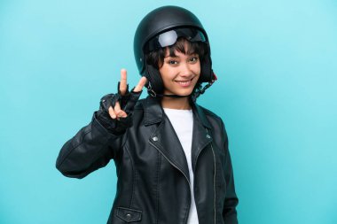 Mavi arka planda izole edilmiş motosiklet kasklı genç Arjantinli kadın gülümsüyor ve zafer işareti gösteriyor.