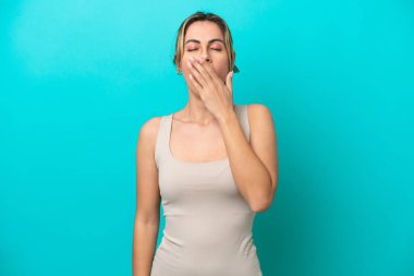 Genç beyaz kadın mavi arka planda izole edilmiş esniyor ve geniş bir ağzı eliyle kapatıyor.