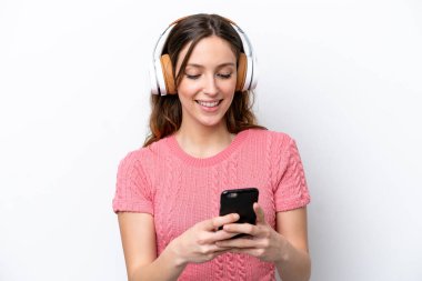 Beyaz arka planda izole edilmiş genç beyaz kadın müzik dinliyor ve cep telefonu arıyor.