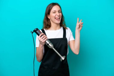 Beyaz tenli genç aşçı kadın mavi arka planda izole edilmiş el blenderi kullanıyor gülümsüyor ve zafer işareti gösteriyor.