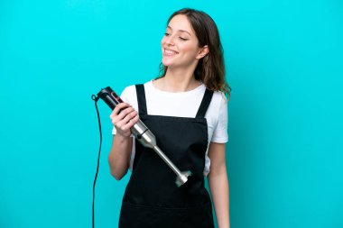 Beyaz tenli genç aşçı kadın mavi arka planda izole edilmiş el blenderi kullanıyor. Yan tarafa bakıyor ve gülümsüyor.