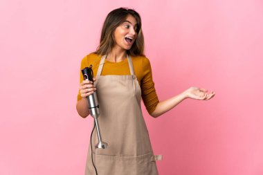 Genç aşçı kadın, el blenderi kullanıyor. Pembe arka planda saklanmış, sürpriz bir ifadeyle yan tarafa bakarken.