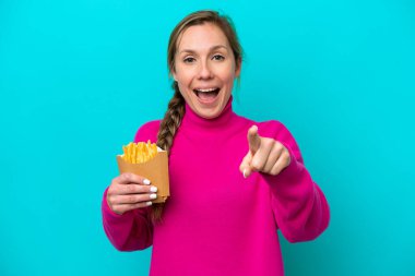 Elinde kızarmış cips tutan genç beyaz kadın mavi arka planda izole edilmiş şaşırmış ve ön tarafı işaret ediyor.
