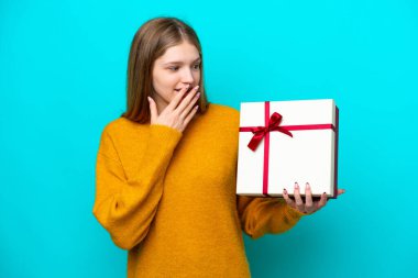 Teenager Russian girl holding a gift isolated on blue background with surprise and shocked facial expression